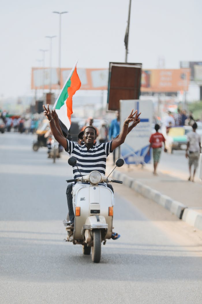 service-02 Enthusiastic man rides scooter waving Sudanese flag in a vibrant city street, expressing joy and freedom.