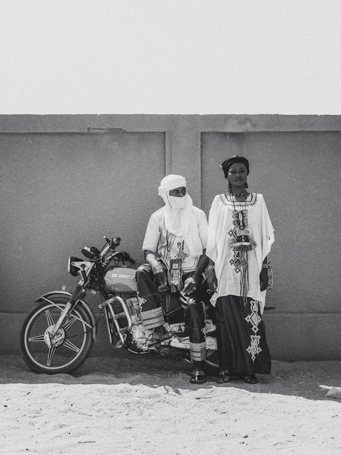 service-06 Black and white photo of a man and woman in traditional attire, sitting and standing by a motorbike in Niger.